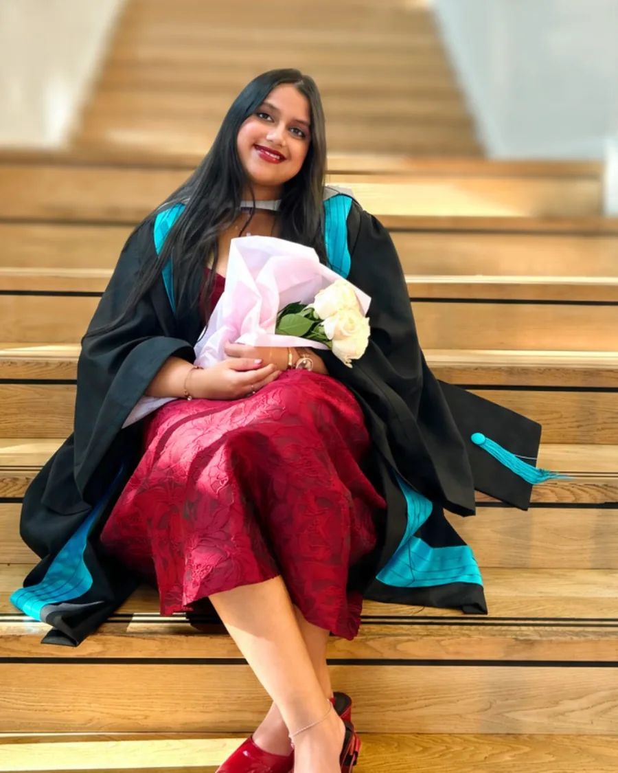 Vibha in her graduation gown and a red dress, sitting on a wooden staircase with white roses.