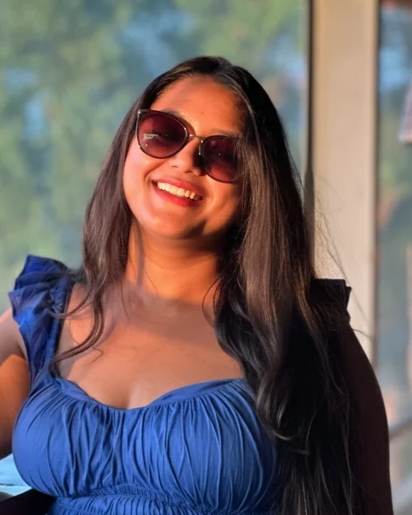 Vibha smiling in sunglasses and a blue dress, in warm evening light.