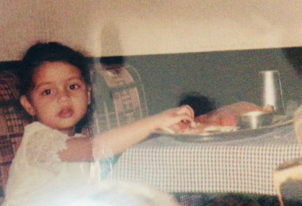 Vibha as a toddler, hand on a plate of dosa, looking up at the camera mid-bite.