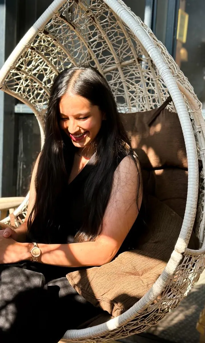 A woman in a wicker swing chair laughing, looking down, in dappled sun.