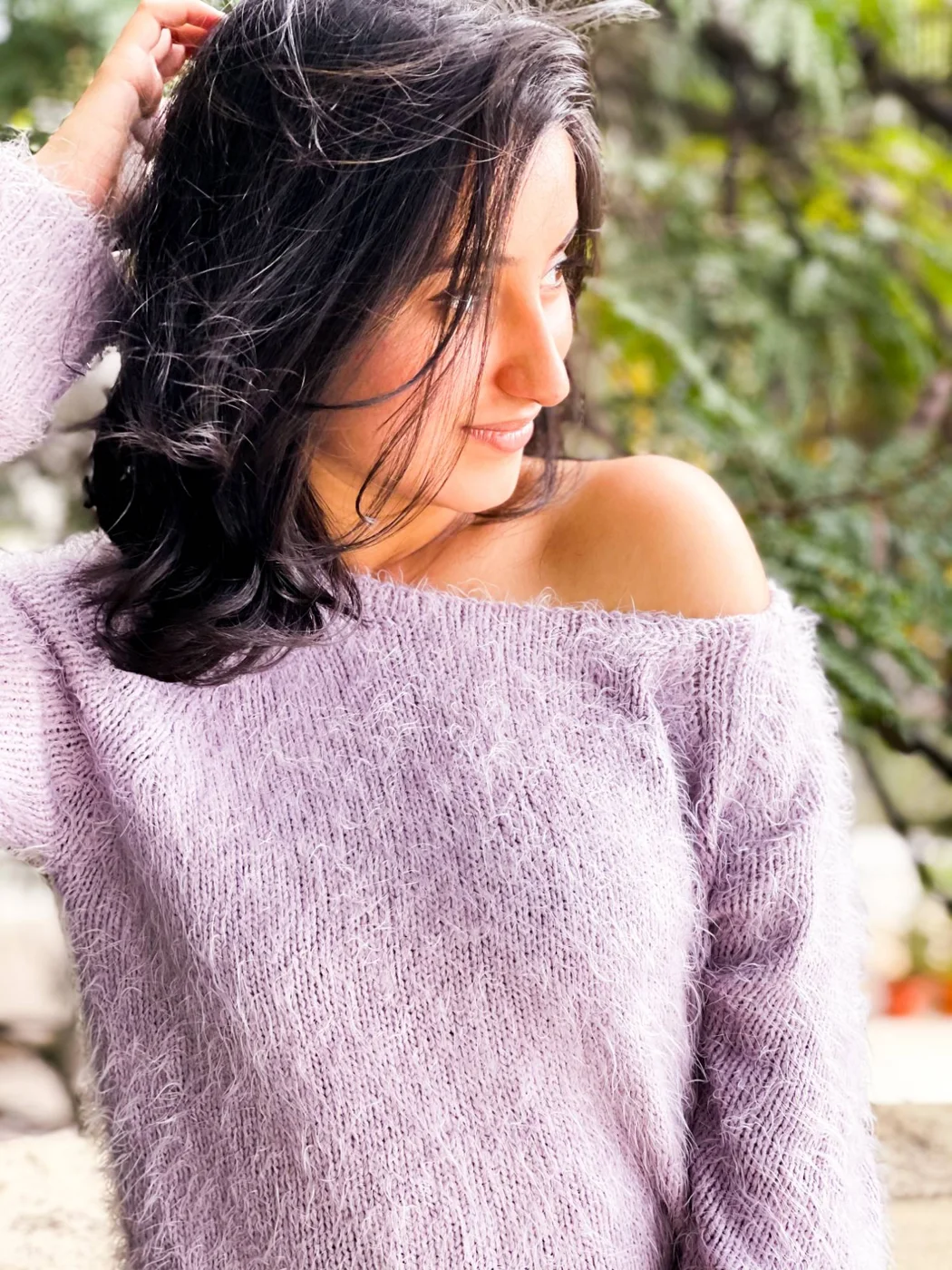 A woman in a fluffy lilac jumper glancing over her bare shoulder, hand in her hair, greenery behind.