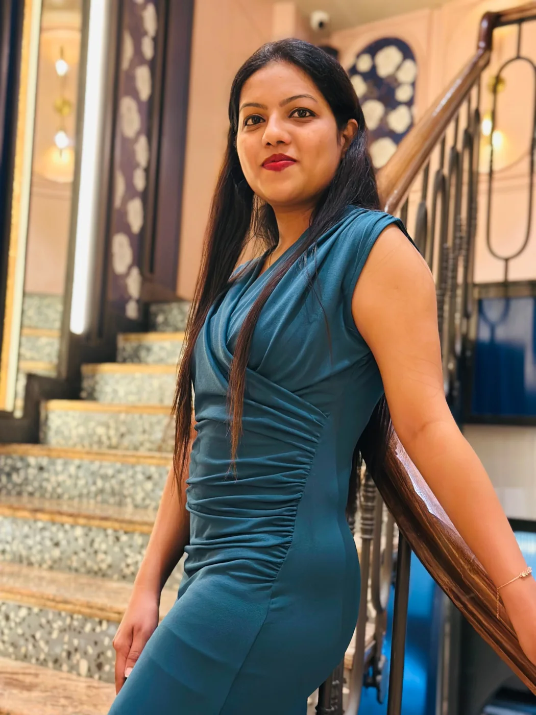A woman in a teal dress leaning on a wooden banister on a tiled staircase.