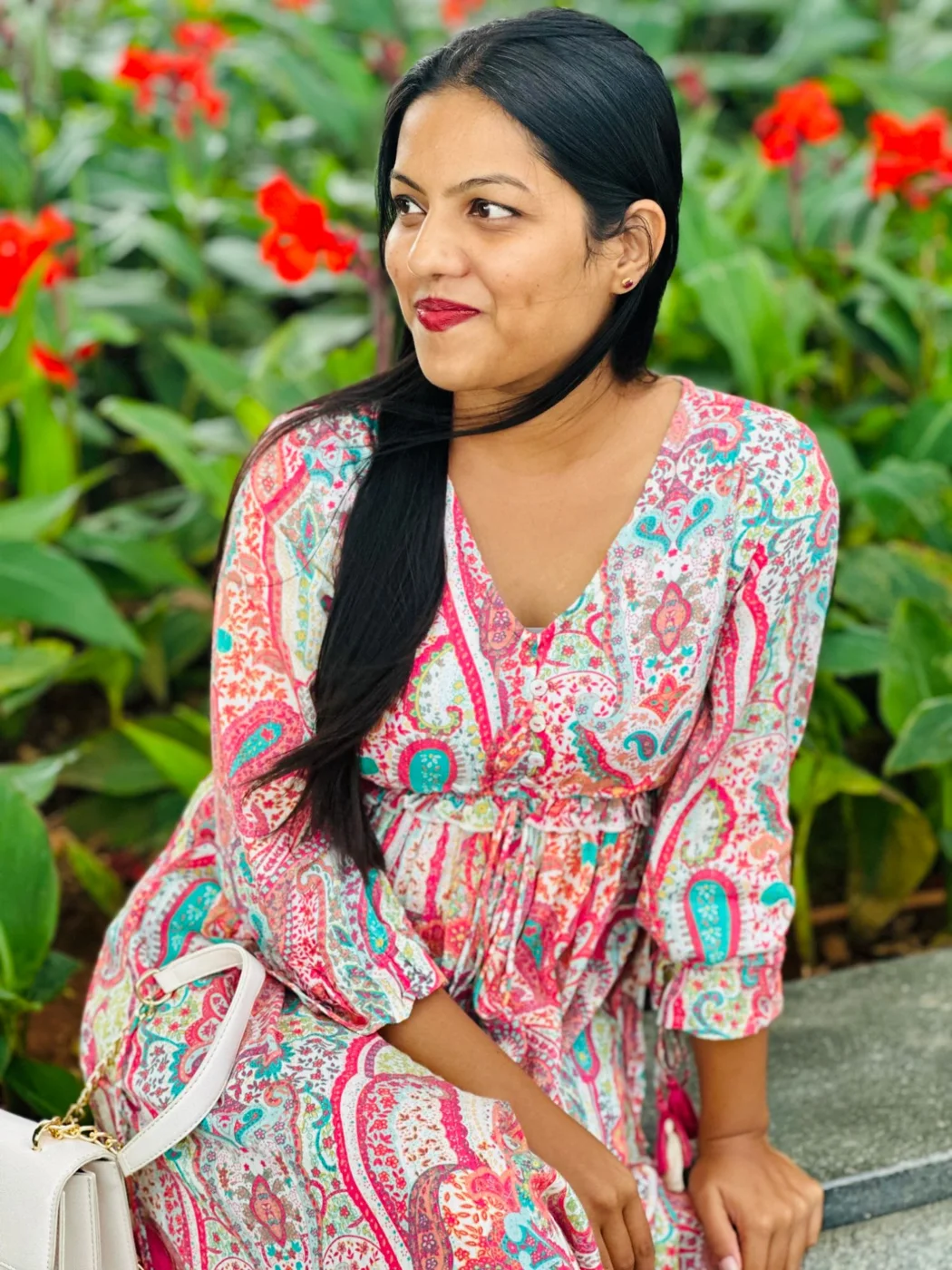A woman in a pink paisley dress sitting by red flowers, glancing to one side.