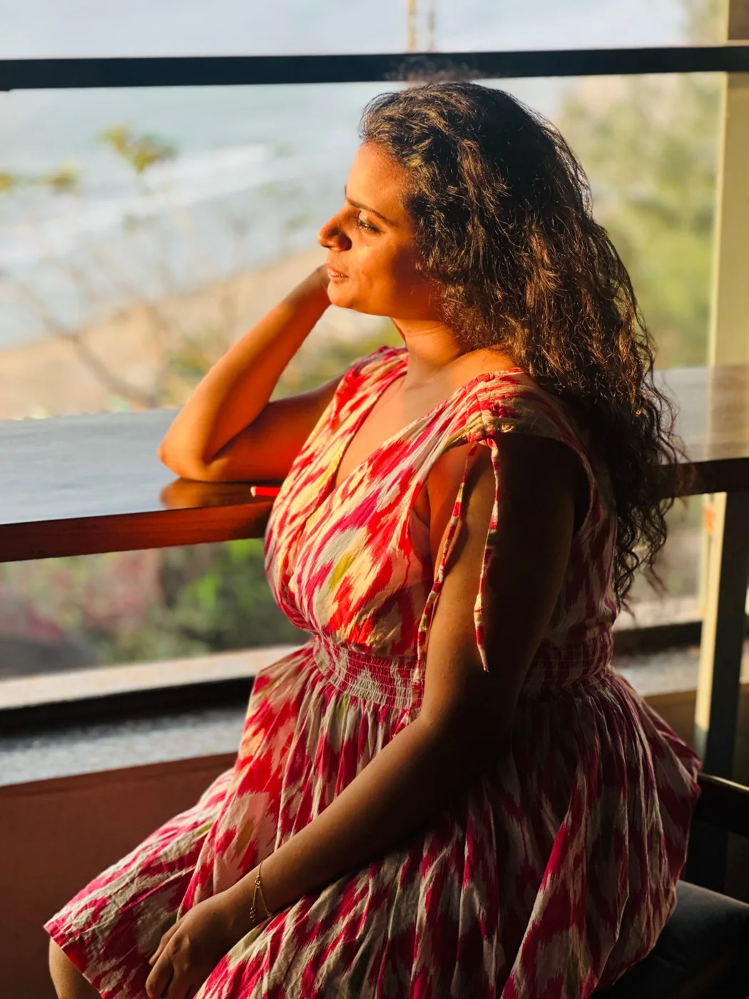 A woman in a pink ikat dress resting her elbow on a ledge, looking out at the sea in warm light.