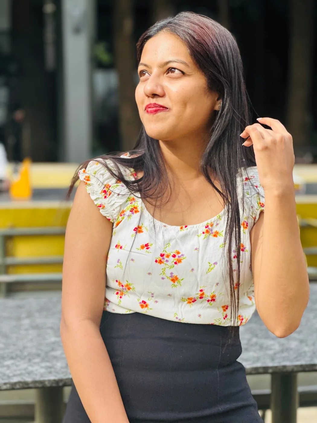 A woman in a floral top and black skirt looking away, one hand in her hair.