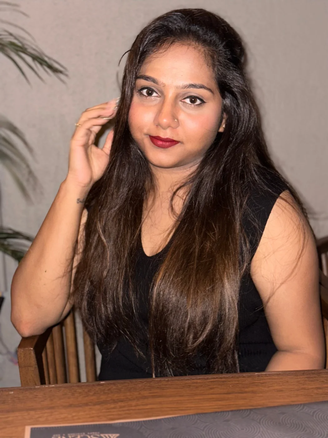 A woman with long hair in a black dress seated at a wooden table, touching her hair.