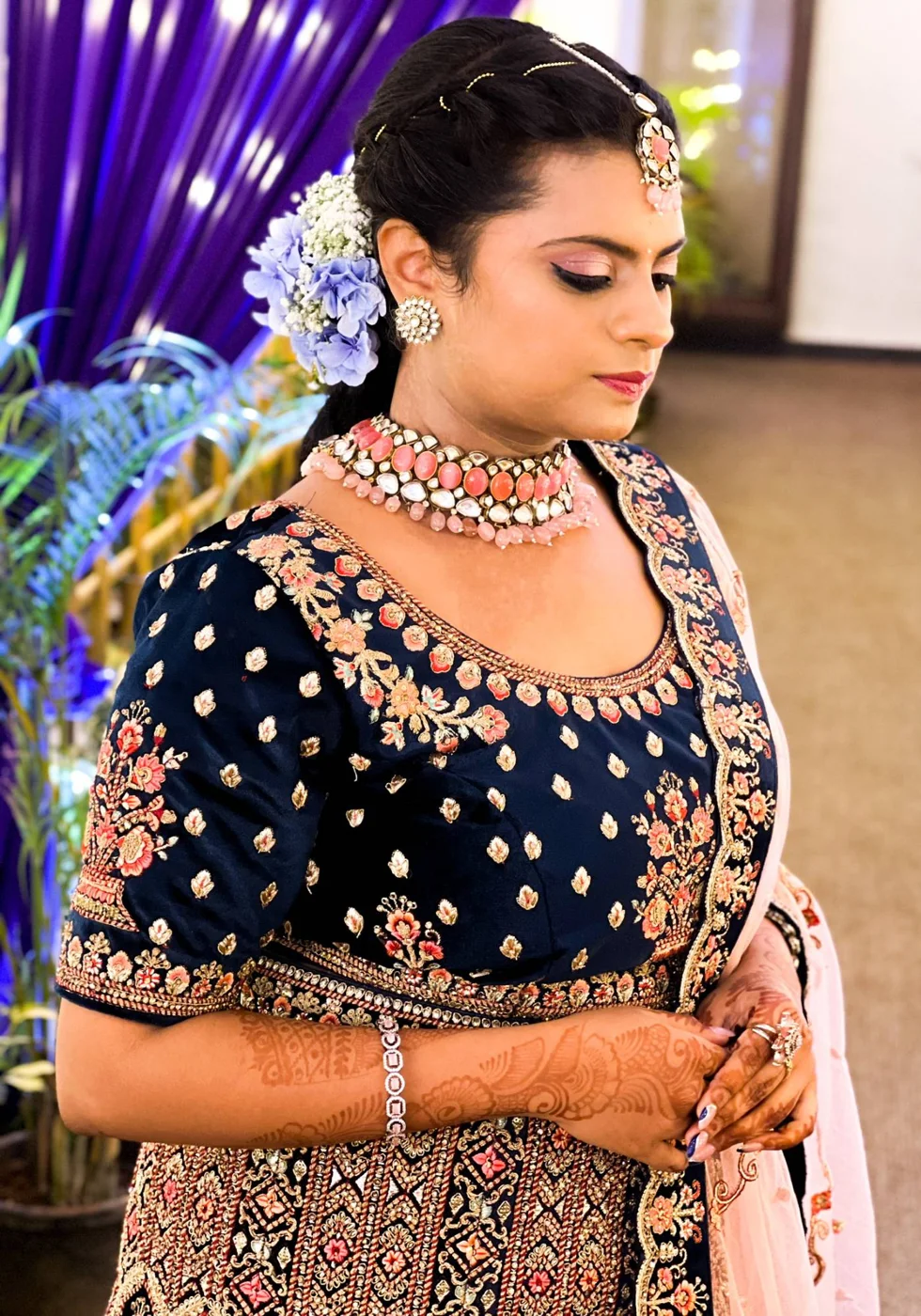 A bride in a navy embroidered lehenga and pink choker, looking down, flowers in her hair.