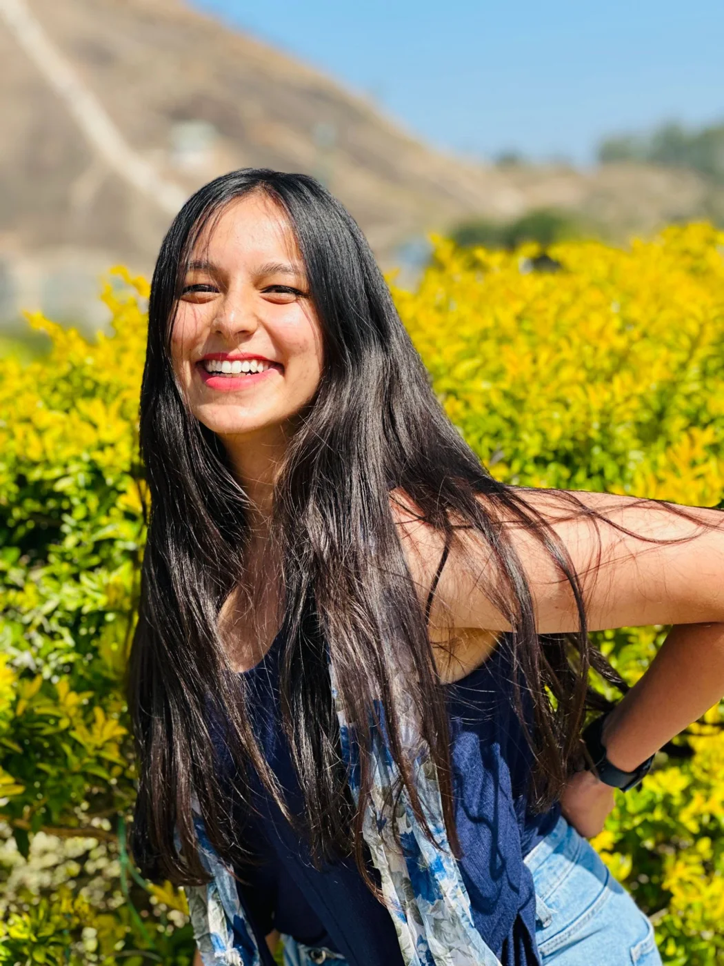 A woman laughing in front of bright yellow shrubs, long hair blowing across her.